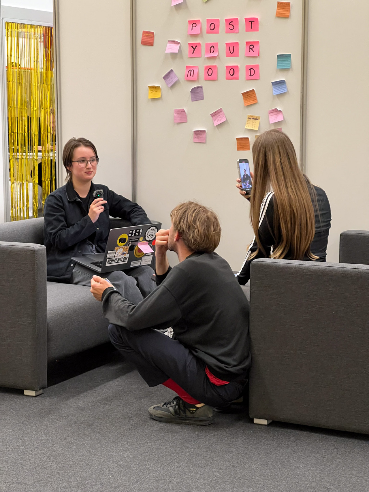 Cast students are sitting on sofas and record a video using a microphone and a phone camera. Post-its on the wall say: "Post Your Mood". Credits: Nadia Holdener.