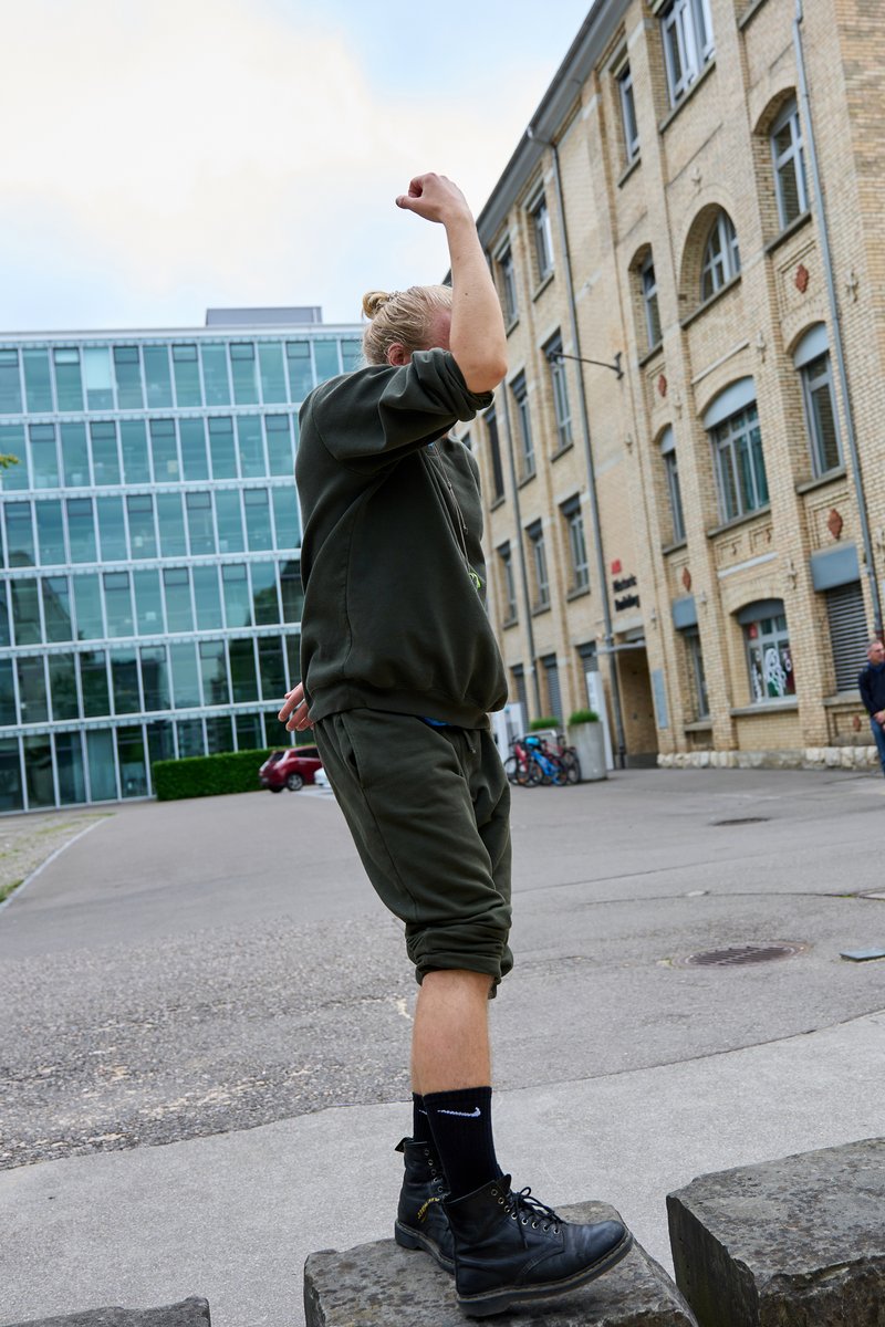 Performance: untitled dance (47.4122,8.54016), 2023, MFO-Park, Zürich  Während der Performance, in grünen Jogginghosen auf einem Felsen stehend in einer Position, die das Gesicht verdeckt; im Hintergrund sind Gebäude zu sehen. Bild: Maria Cheilopoulou und TanzLobby