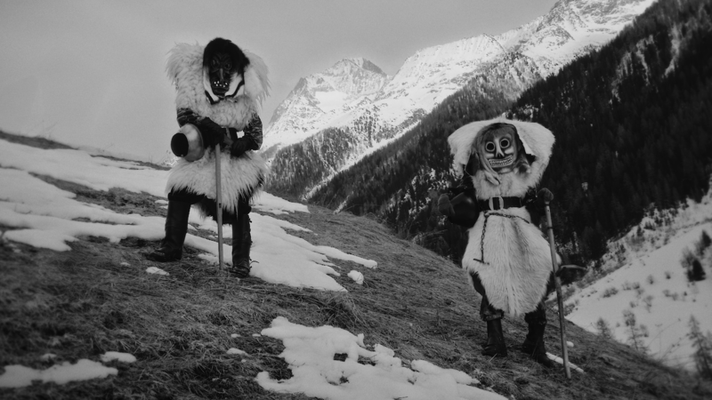 Berglandschaft mit Schnee, der den Boden nicht durchgehend bedeckt. Zwei Personen in Fellen, Masken und mit Kuhglocken. © ZHdK.