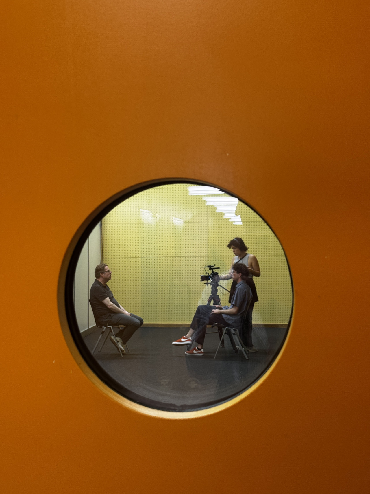 Durch ein kleines Fenster sieht man in ein Aufnahmestudio, in dem zwei Cast-Studierende ein Interview mit einer Person aufnehmen. Credits: Adrian Graf.