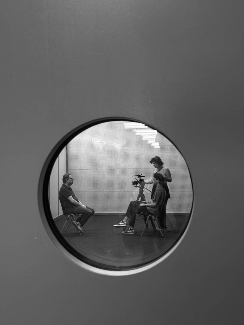 Durch ein kleines Fenster sieht man in ein Aufnahmestudio, in dem zwei Cast-Studierende ein Interview mit einer Person aufnehmen. Credits: Adrian Graf.