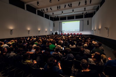 A big hall at the Museum für Gestaltung; Many people in the audience. On stage there are five people, the screen behind them says