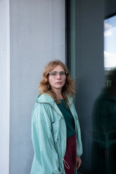 Photograph of a woman wearing glasses and a turquoise jacket standing next to a modern glass façade. Dark windows reflect her silhouette and a bright outdoor area, highlighting the surrounding architecture.