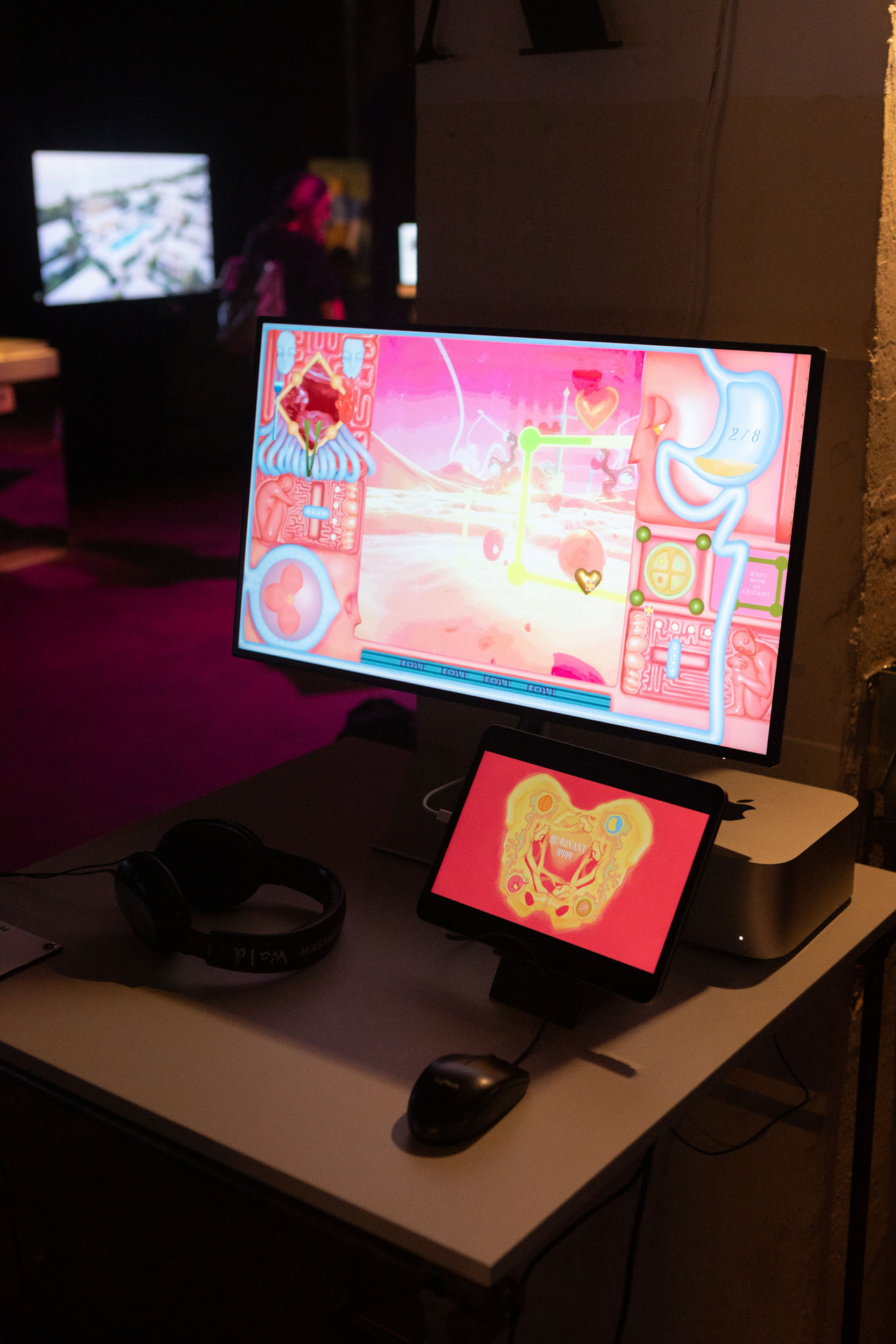 Ein Computerscreen zeigt eine rosa Landschaft und Spielerinterface. Ein iPad zeigt ein Making-Of.