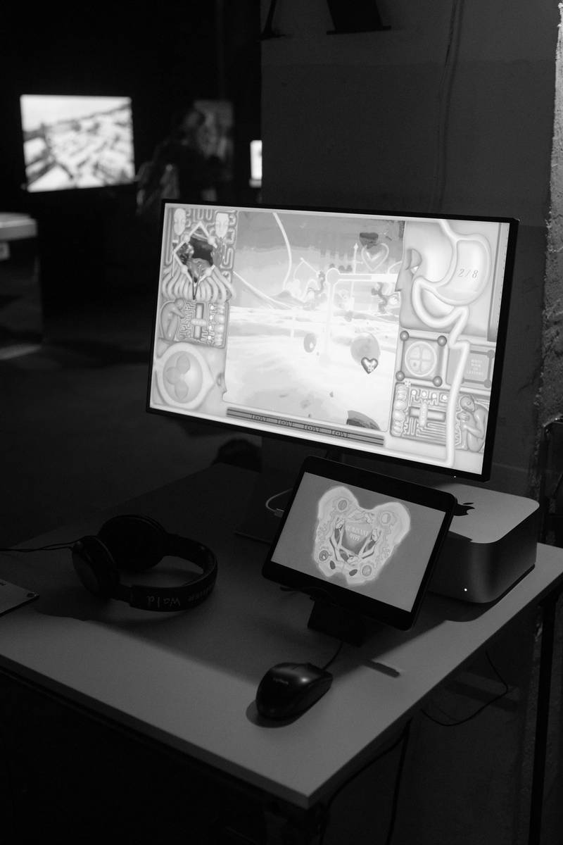 Ein Computerscreen zeigt eine rosa Landschaft und Spielerinterface. Ein iPad zeigt ein Making-Of.